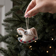 A hand hangs a small embroidered rocking horse ornament from a string in front of a decorated Christmas tree with warm lights.