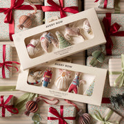 Two boxed sets of Avery Row Christmas tree decorations arranged on top of wrapped gift boxes with red ribbons and ornaments. The boxes contain small fabric holiday ornaments shaped like a bear, bird, tree, and other festive forms, visible through clear window packaging.