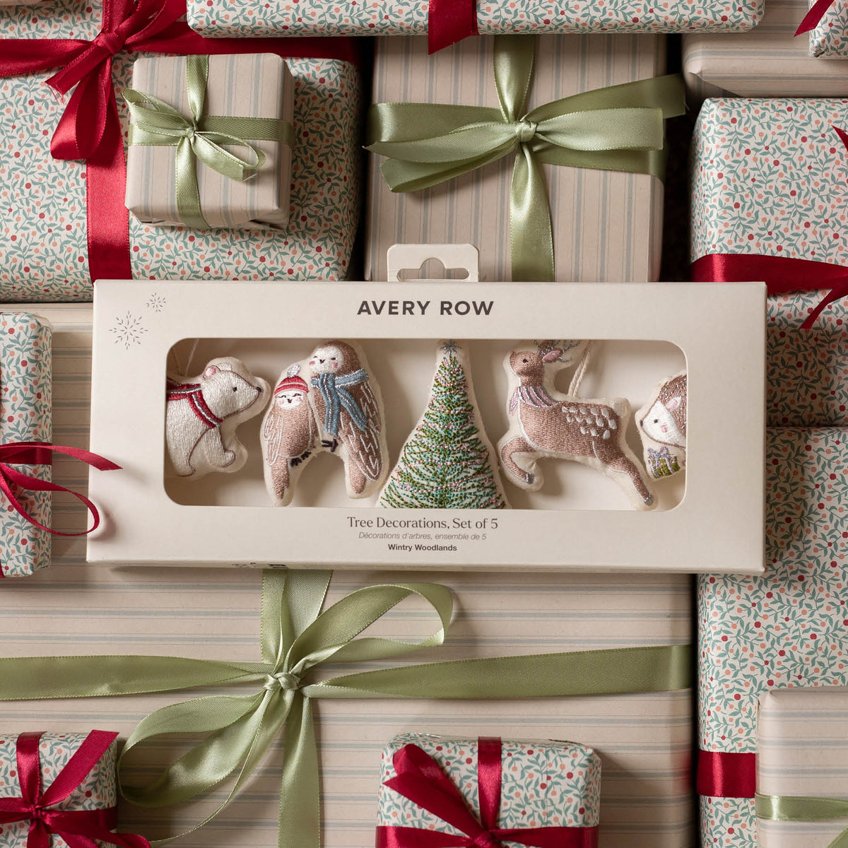 Avery Row Christmas tree decorations set of 5 in a cream-colored gift box, displayed on top of wrapped gift boxes with red ribbons and green satin bows.
