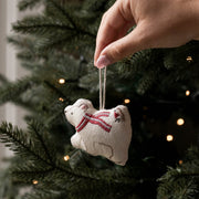A hand hangs a small beige fabric ornament shaped like a polar bear wearing a red striped scarf from a dark green Christmas tree with warm string lights.