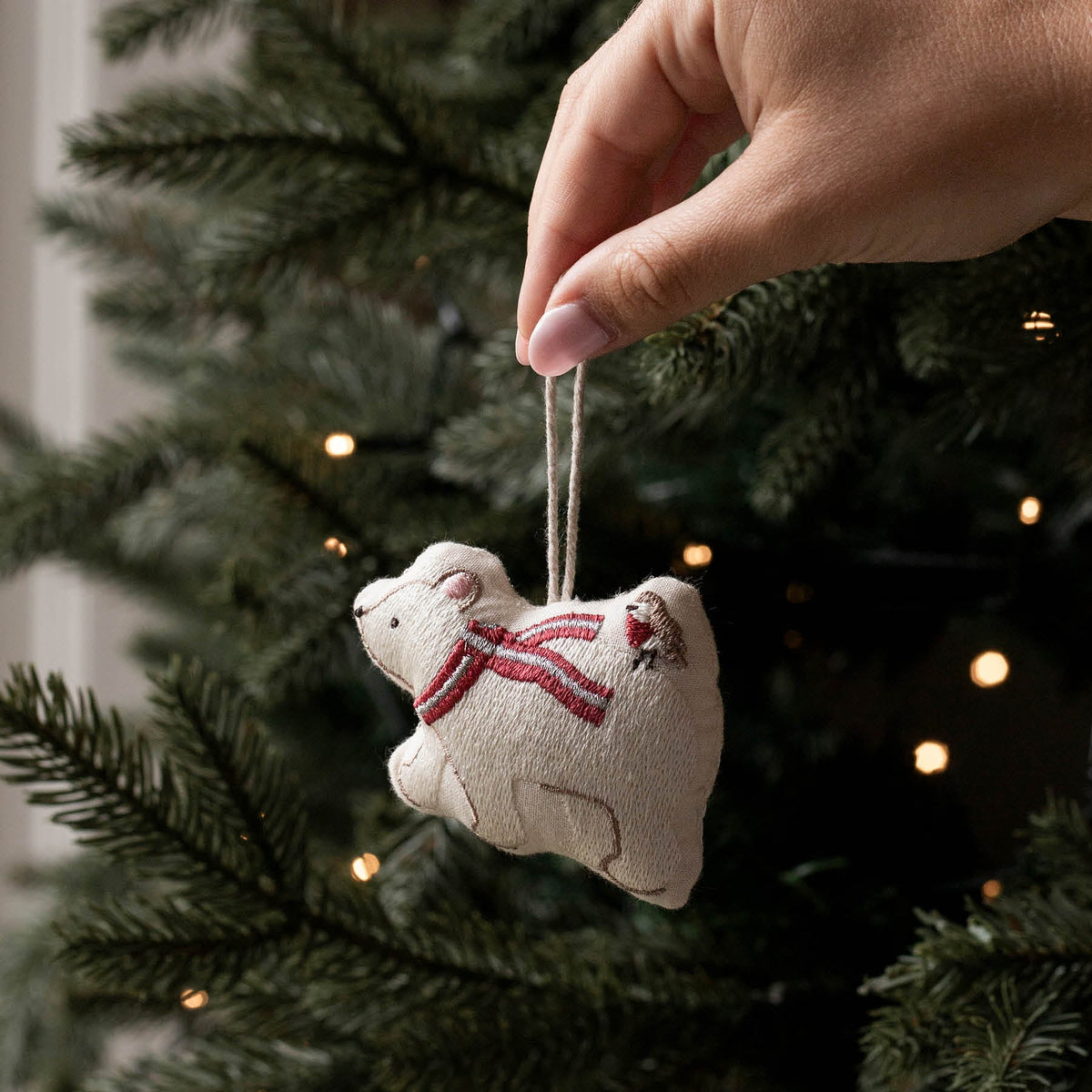 A hand hangs a small beige fabric ornament shaped like a polar bear wearing a red striped scarf from a dark green Christmas tree with warm string lights.