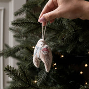 A hand hangs a small embroidered fabric ornament shaped like a pair of owls on a Christmas tree. The ornament is beige with white embroidery and a red scarf, with the tree branches and warm string lights blurred in the background.