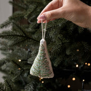 A hand holds a small triangular fabric Christmas tree ornament with green and white embroidered branches, hanging in front of a real Christmas tree with warm string lights.
