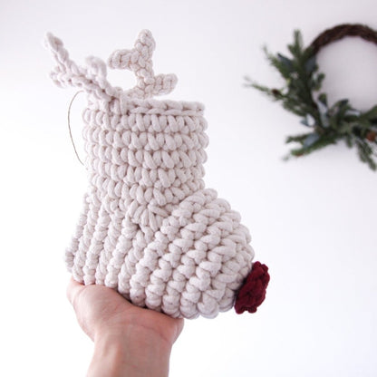 Crocheted reindeer Christmas stocking with antlers and a red nose held by a hand with a wreath in the background.