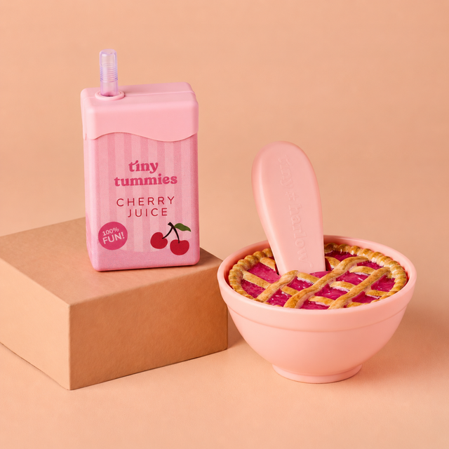 A pink carton-shaped product labeled 'tiny tummies CHERRY JUICE' stands on a brown box beside a pink bowl with a lattice-top lid and a matching pink spoon, all on a peach studio background.