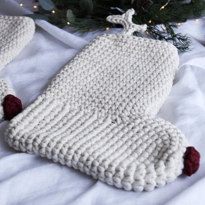Knitted white stocking with red nose and antlers on a white surface with Christmas decorations.