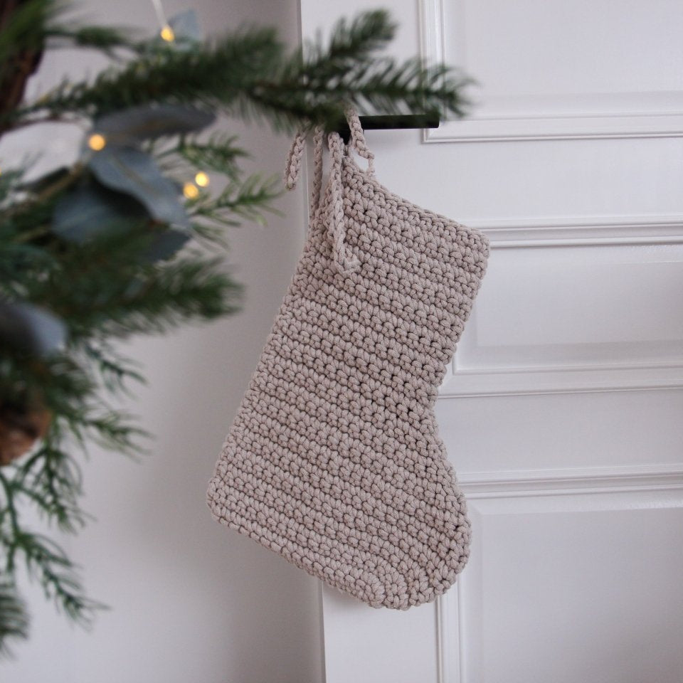 Knitted beige stocking hanging on a white wall with a Christmas tree in the background.