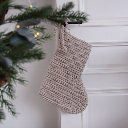 Knitted beige stocking hanging on a white wall with a Christmas tree in the background.