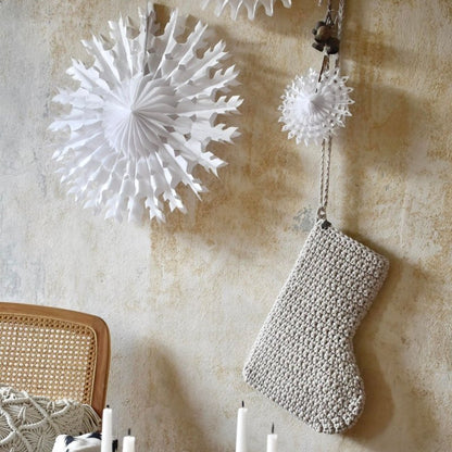 Decorative white paper snowflakes and a textured stocking hanging on a beige wall with candles and greenery.