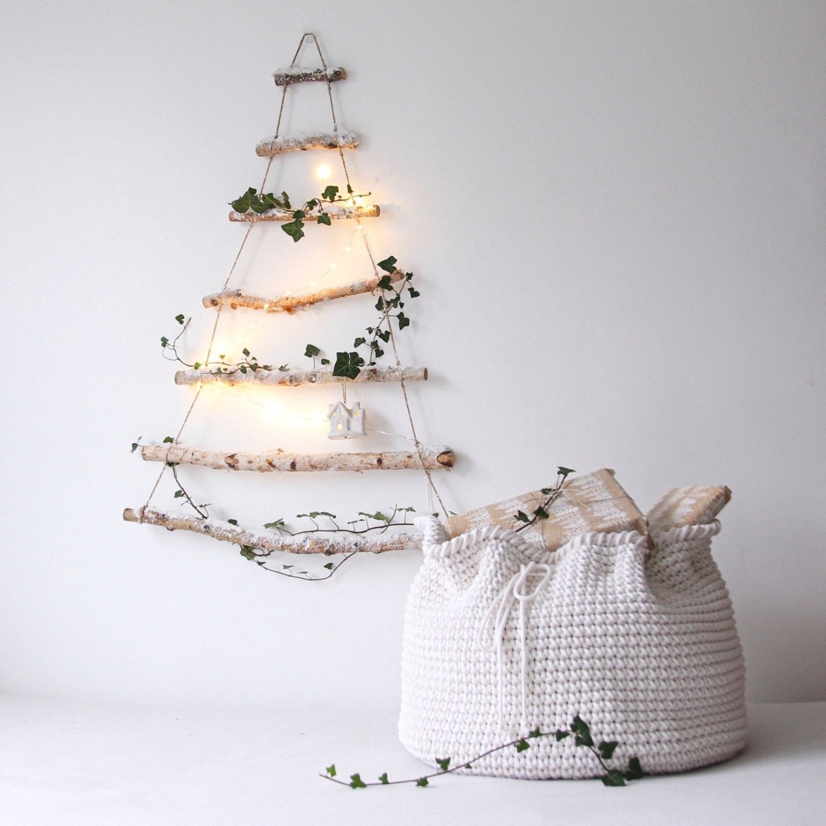 Decorative wall art made of wooden twigs with lights and greenery, next to a white crochet present sack on a light background.