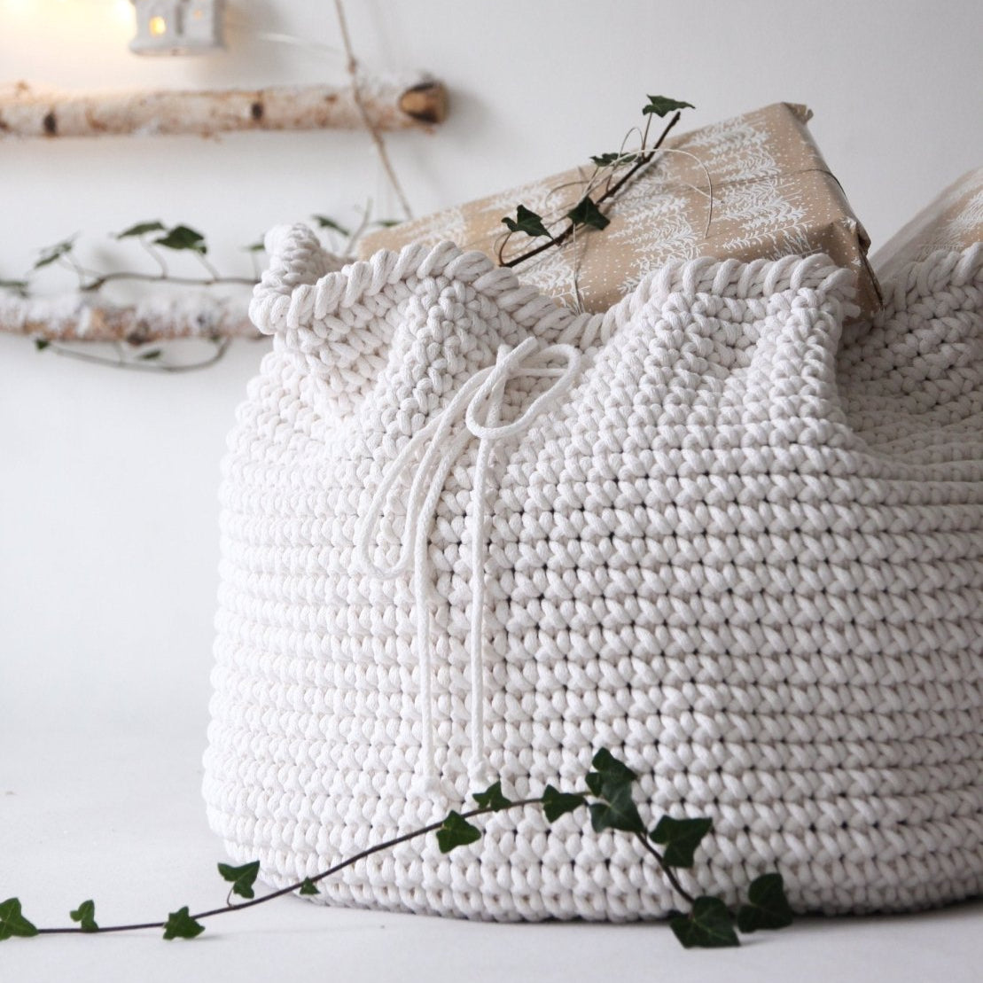 White knitted present sack with drawstring on a white surface with green leaves.