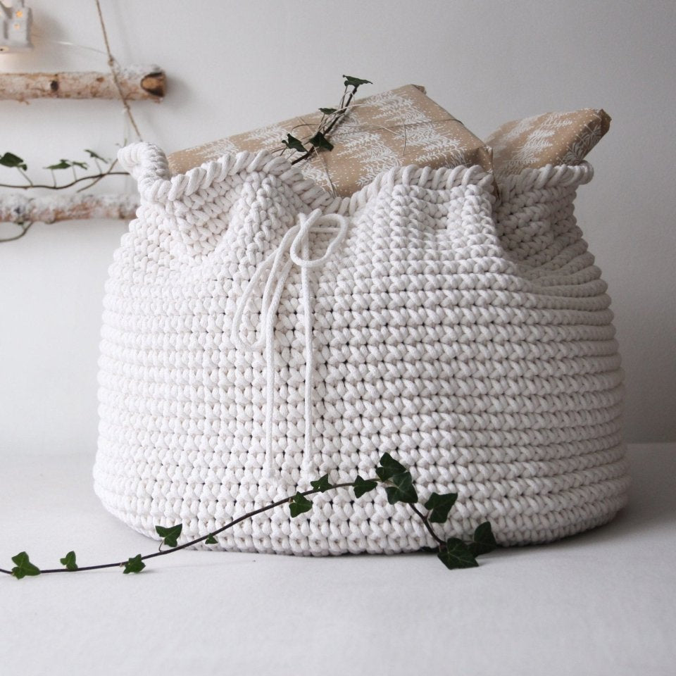 White crocheted present sack with a drawstring on a white surface with decorative branches.