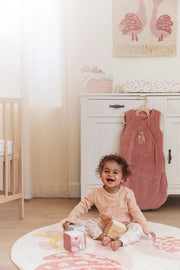 A baby sitting on a round rug in a nursery, wearing a pink long-sleeve top and patterned trousers. A dusty pink swaddle blanket hangs on a wooden hanger on a white dresser behind them.
