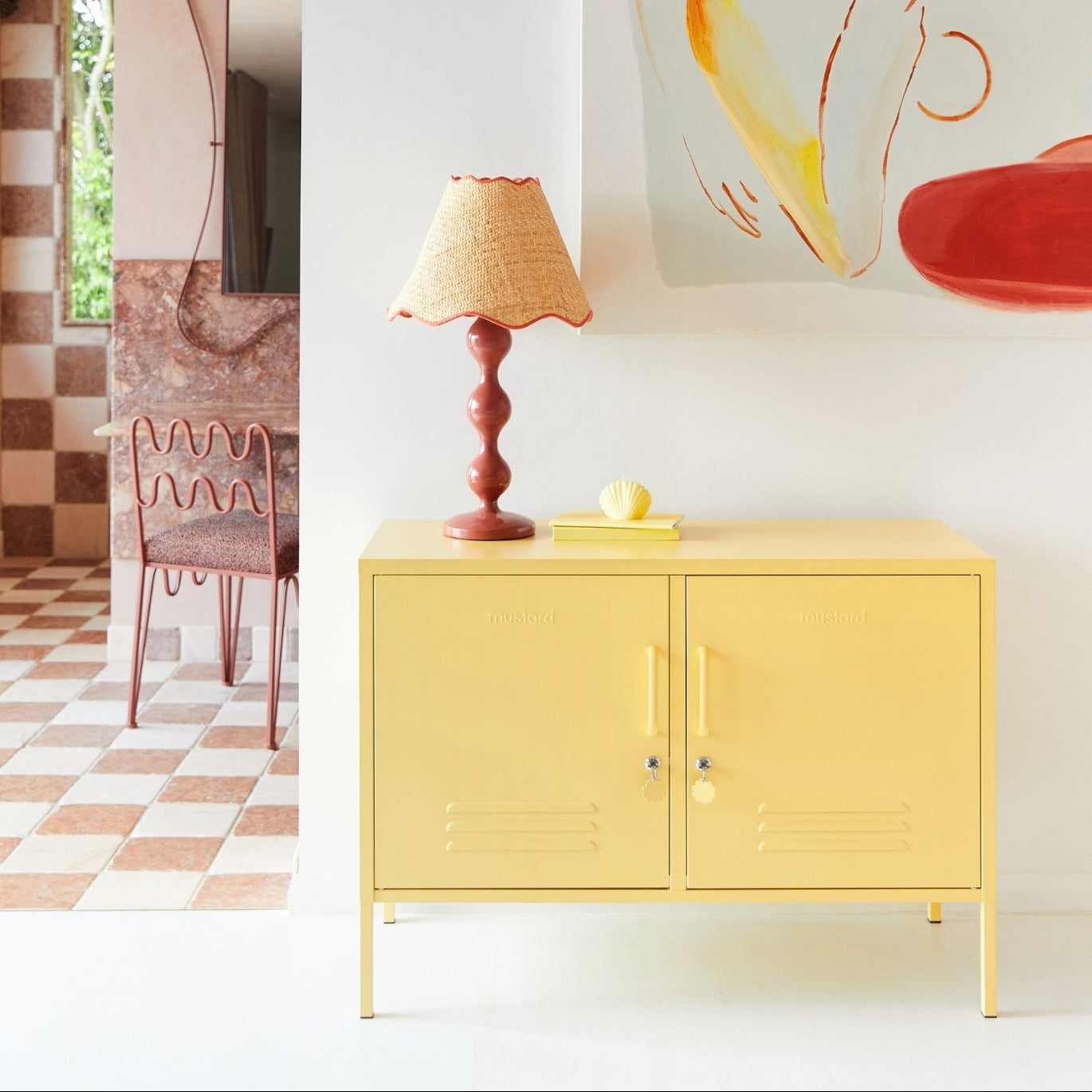 Yellow cabinet with a lamp on top in a room with a checkered floor and colorful wall art.