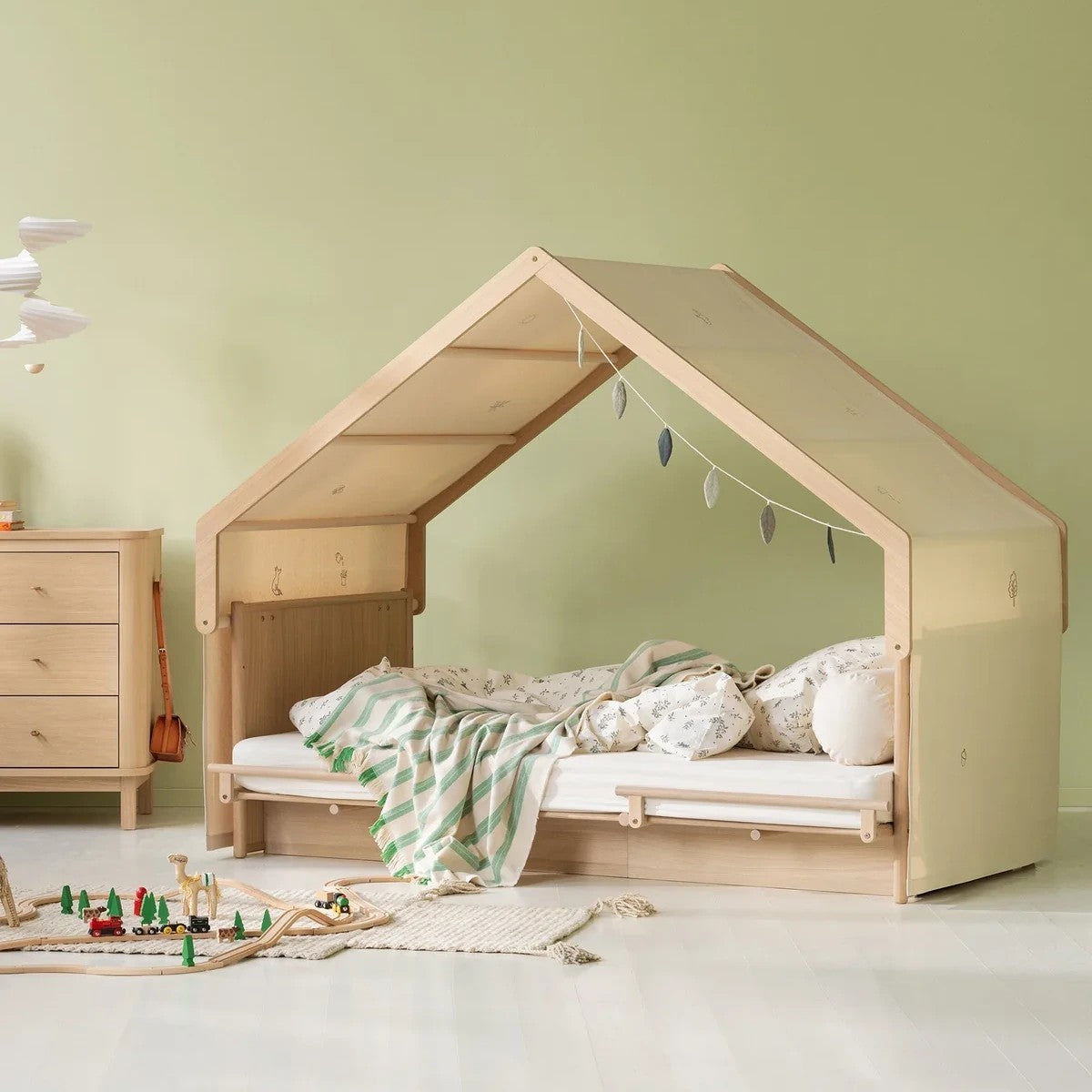 A house-shaped wooden children's bed with a slanted roof, styled in a nursery with a green wall, white floor, a matching wooden dresser, and a toy train on the floor.