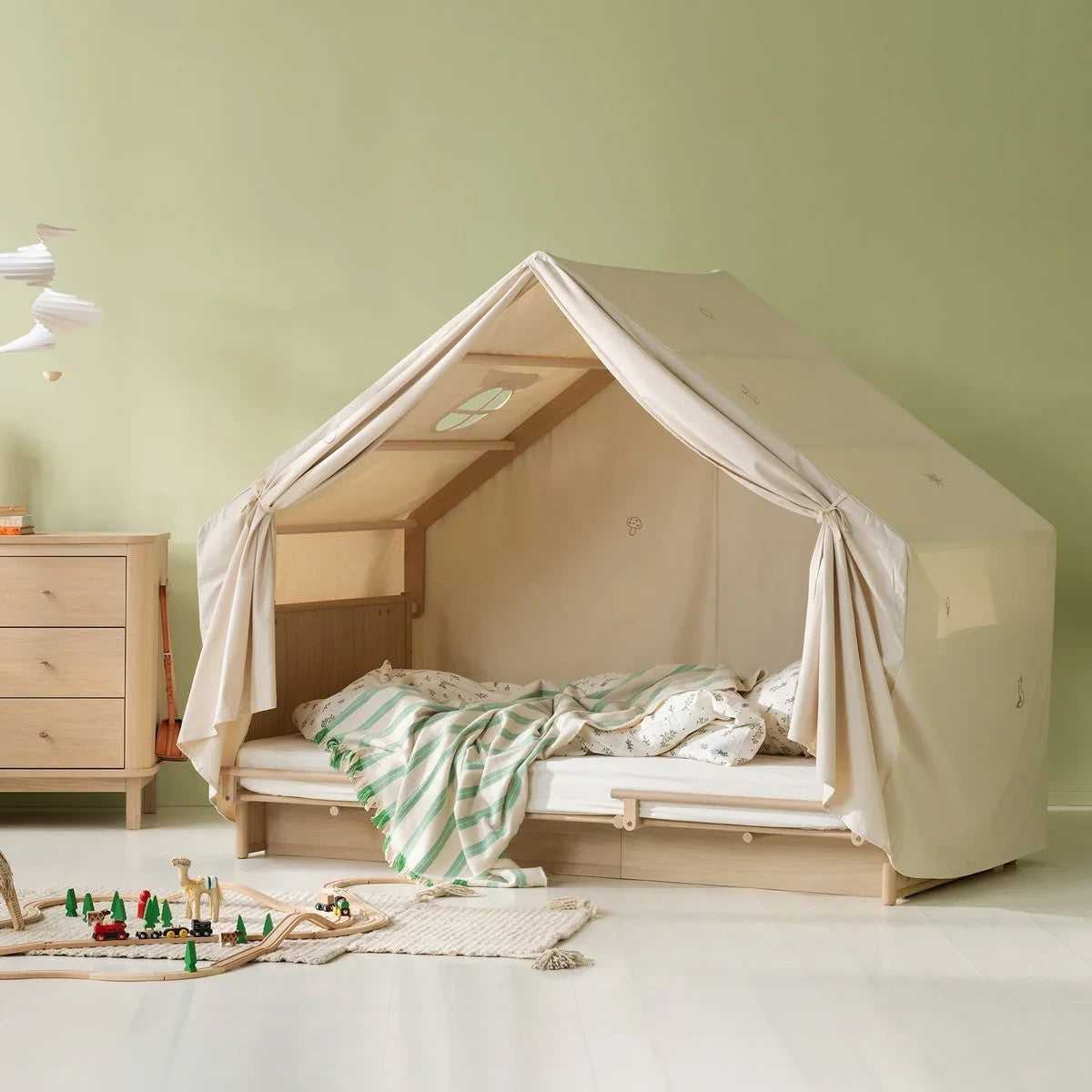 A children's bed styled as a canvas play tent in a nursery, featuring a light wood frame, beige canvas walls, and a green and white striped blanket on a light-colored floor.