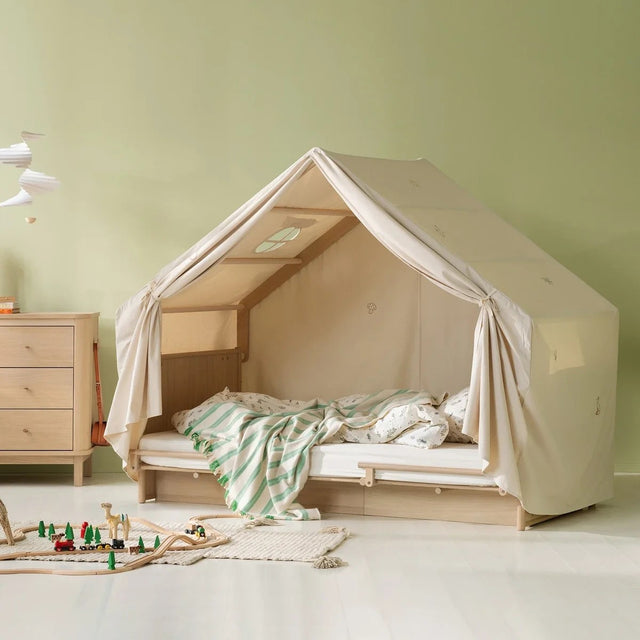 A children's bed styled as a canvas play tent in a nursery, featuring a light wood frame, beige canvas walls, and a green and white striped blanket on a light-colored floor.