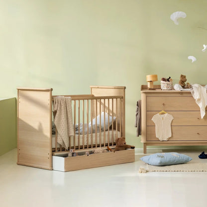 A light natural wood baby crib and matching changing table set in a softly styled nursery with pale green walls, white floor, and small decorative toys and pillows.