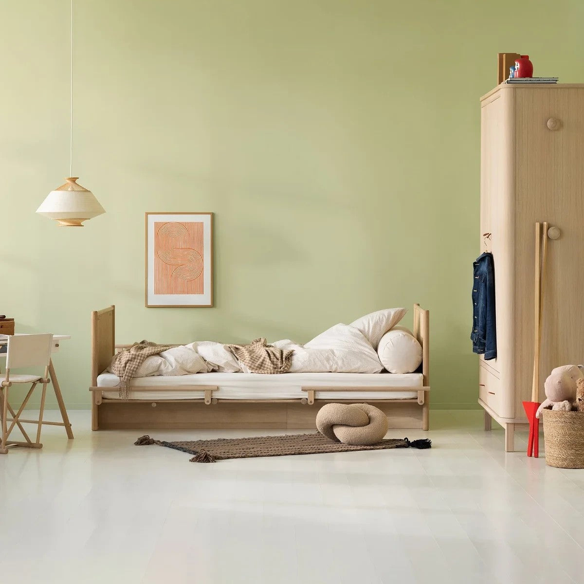 A light wood single bed frame with white bedding and pillows, styled in a minimalist, modern bedroom with a sage green wall, a wooden wardrobe, and a hanging pendant light.