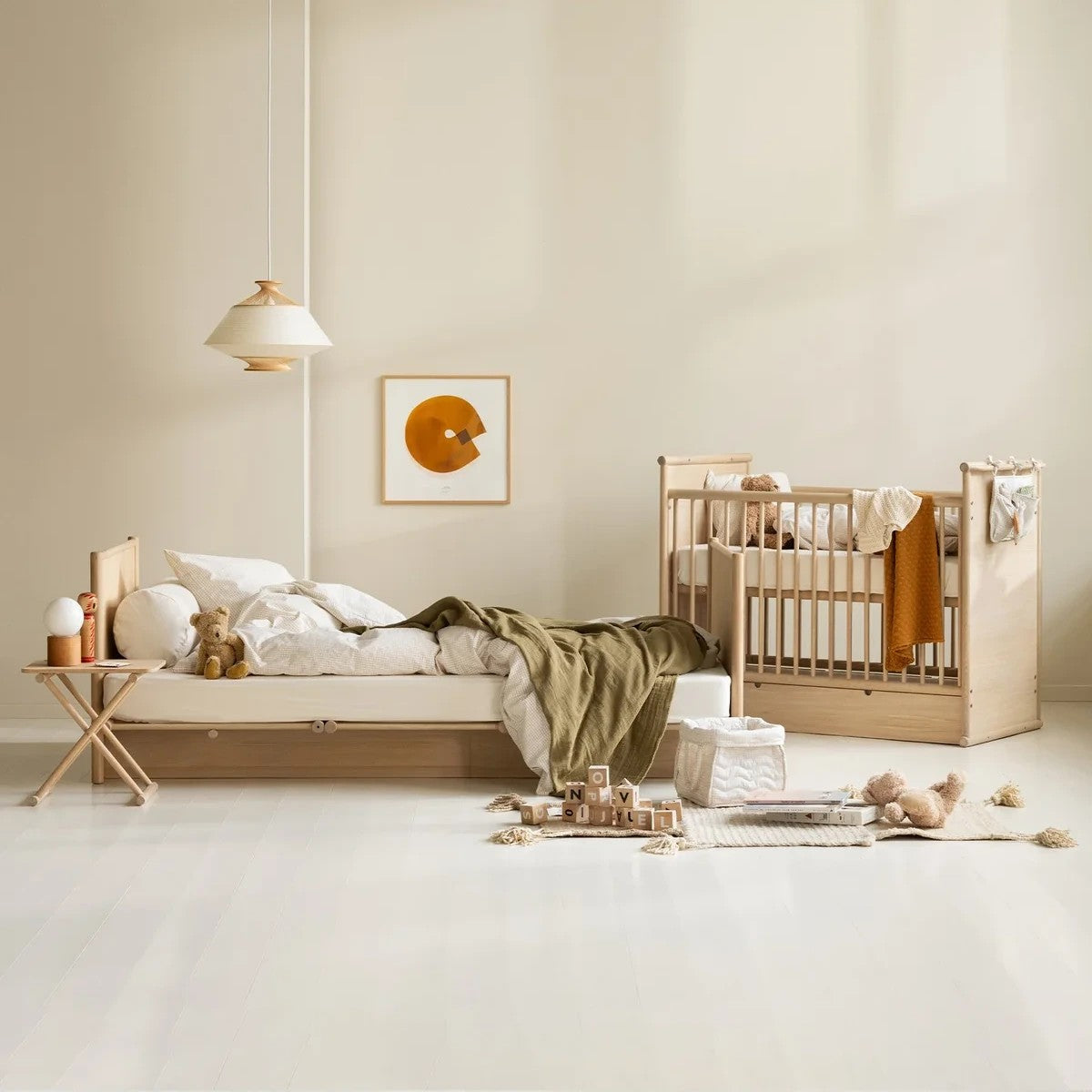 A styled nursery interior showing a light natural wood crib and a matching low bed with white bedding and an olive green throw blanket, set against warm beige walls with a hanging pendant light and framed wall art.
