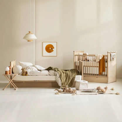 A styled nursery interior showing a light natural wood crib and a matching low bed with white bedding and an olive green throw blanket, set against warm beige walls with a hanging pendant light and framed wall art.