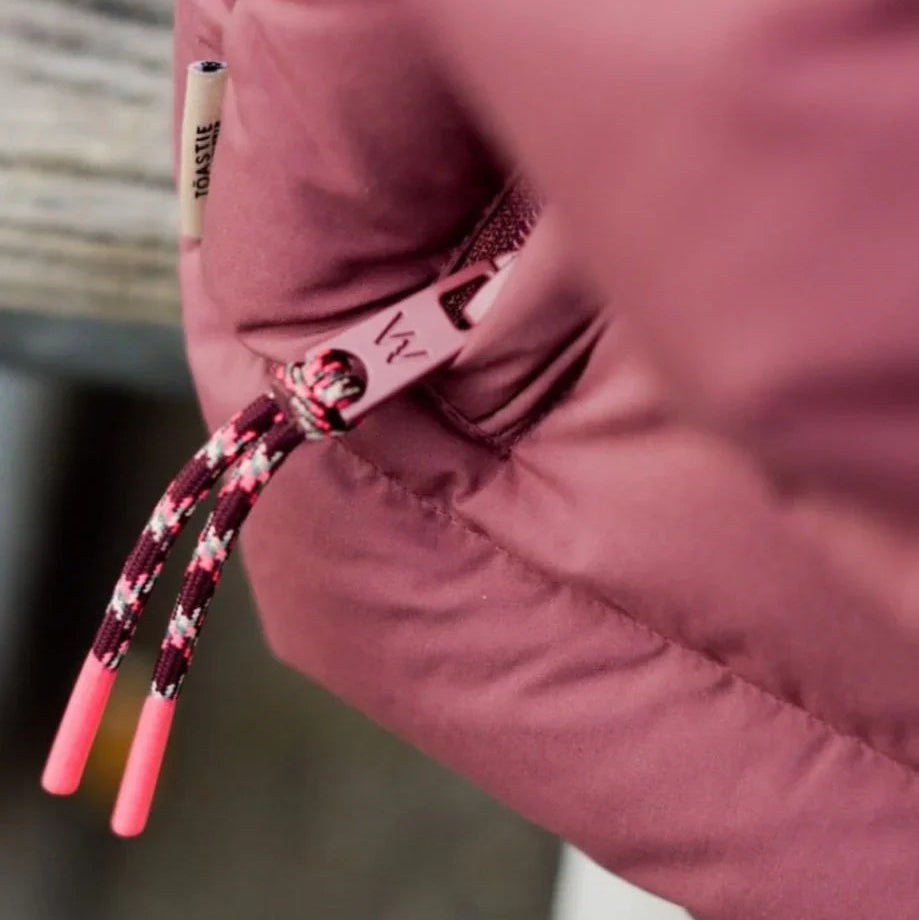 Close-up of a dusty rose puffer jacket with a patterned drawstring and a small white tag that reads 'BADE'.