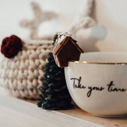 White mug with 'Take your time' text, small Christmas tree, and gingerbread house on a wooden surface.