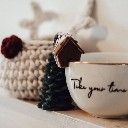 White mug with 'Take your time' text, small Christmas tree, and gingerbread house on a wooden surface.